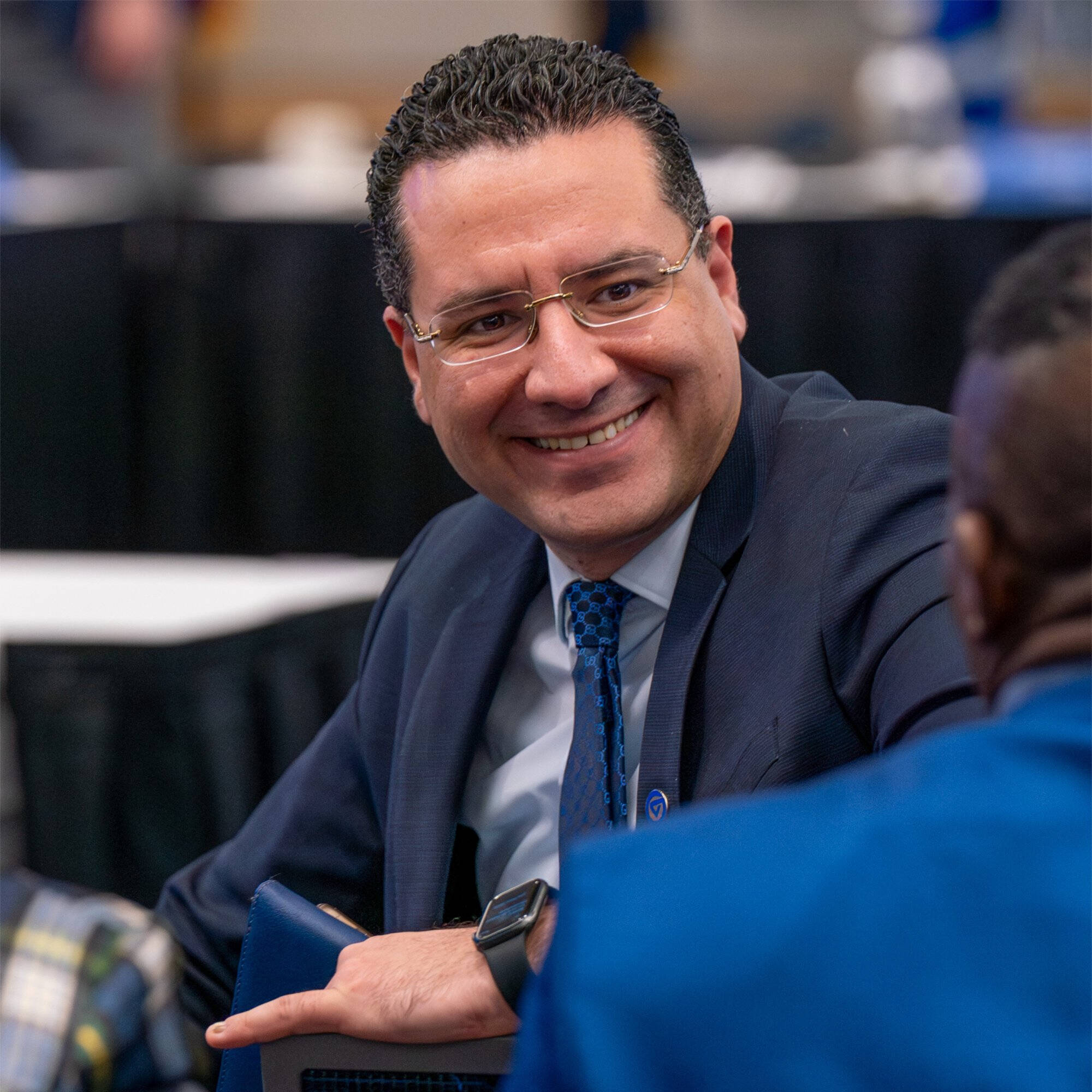 The College Dean in a navy suit, glasses, and blue patterned tie smiles while seated and speaking with others at an event.